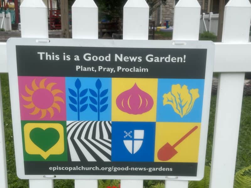 A white fence features a colorful sign for a Good News Garden with symbols of plants, agriculture, and spirituality.