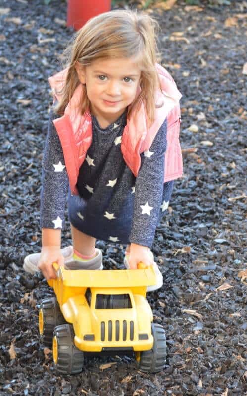 A child with light brown hair plays outside, pushing a yellow toy dump truck on dark mulch while wearing a star-patterned dress and pink vest.
