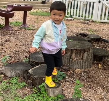 Young child standing on tree stumps in outdoor preschool play area, wearing colorful rain boots and a pastel jacket, engaging in outdoor activities at Trinity Episcopal Preschool.