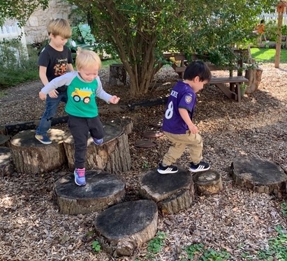 Children exploring outdoor play area at Trinity Episcopal Preschool, balancing on tree stumps in a natural setting.