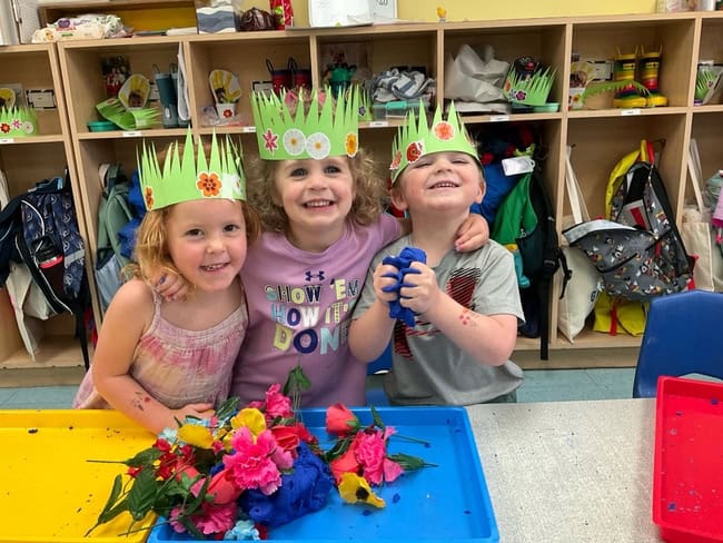 Three happy preschool children wearing paper crown hats with flower decorations, smiling and celebrating in a classroom with colorful cubbies and backpacks in the background.