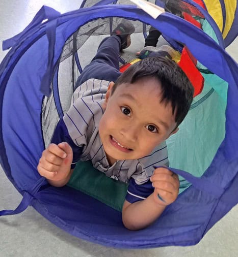 Happy preschool boy crawling through a colorful play tunnel at Trinity Episcopal Preschool, engaging in fun indoor activities that promote early childhood development.